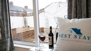 A room with a glass table holding a wine bottle and glass, a pillow with 'NEST STAY' branding, and a view of a brick house through a window. - Bedford Road Garden Apartment (Bedford)
