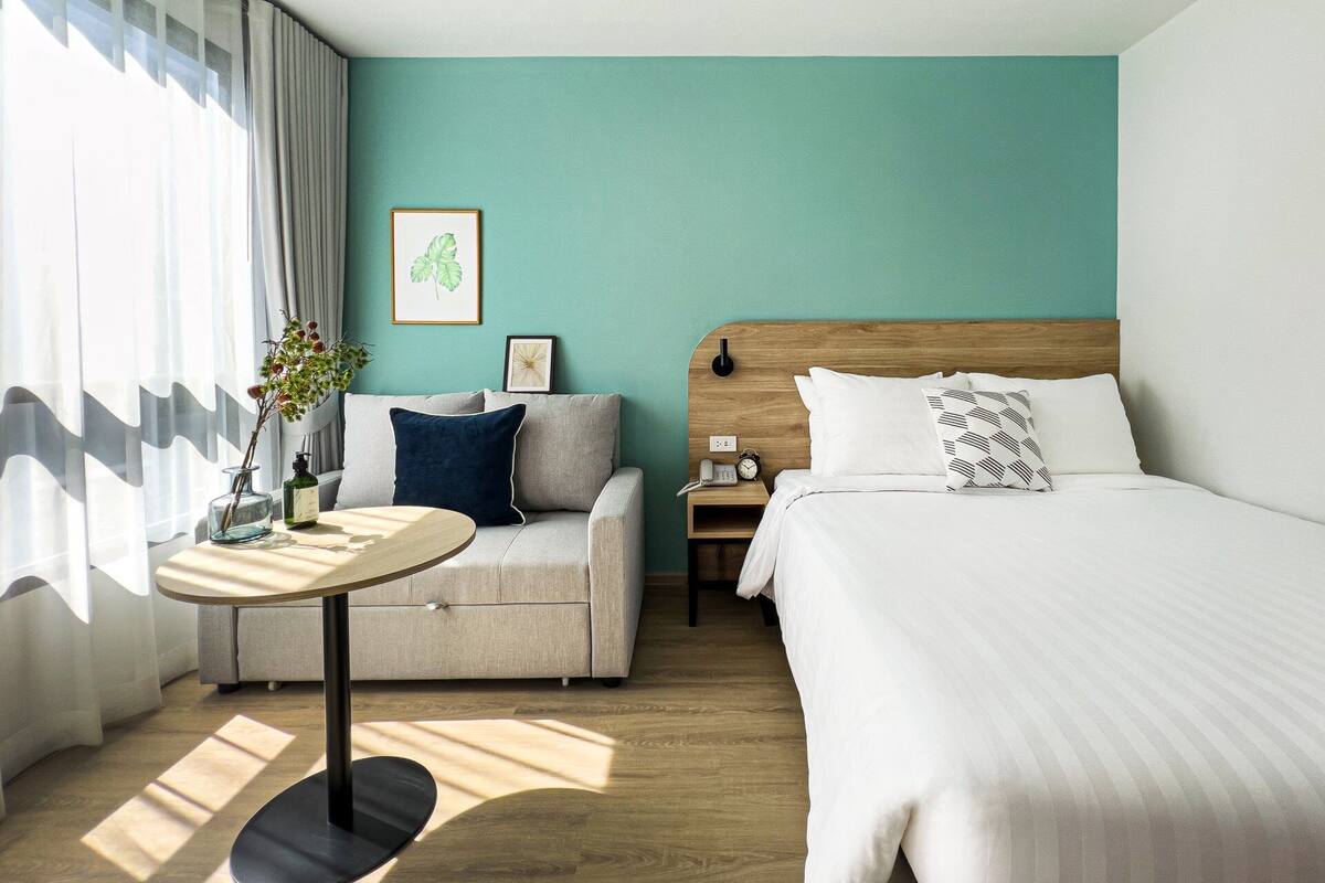 Habitación de hotel con cama, sofá, mesita, mesita de noche y ventana con cortinas.