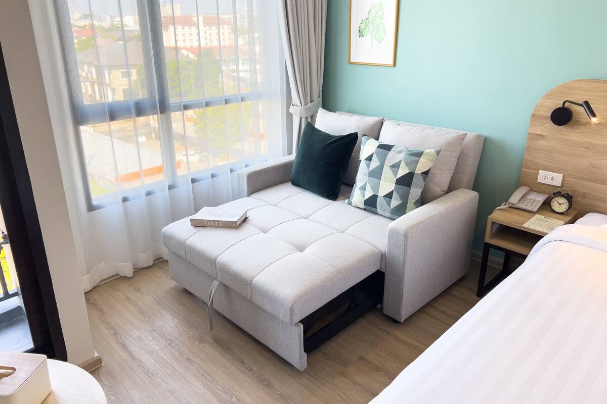 Un dormitorio moderno con sofá cama, mesita de noche con lámpara, reloj y planta enmarcada.