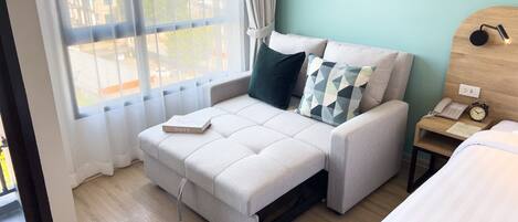 Un dormitorio moderno con sofá cama, mesita de noche con lámpara, reloj y planta enmarcada.