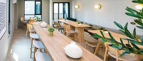 A modern dining area with long wooden tables and chairs, potted plants, and a large window providing natural light.