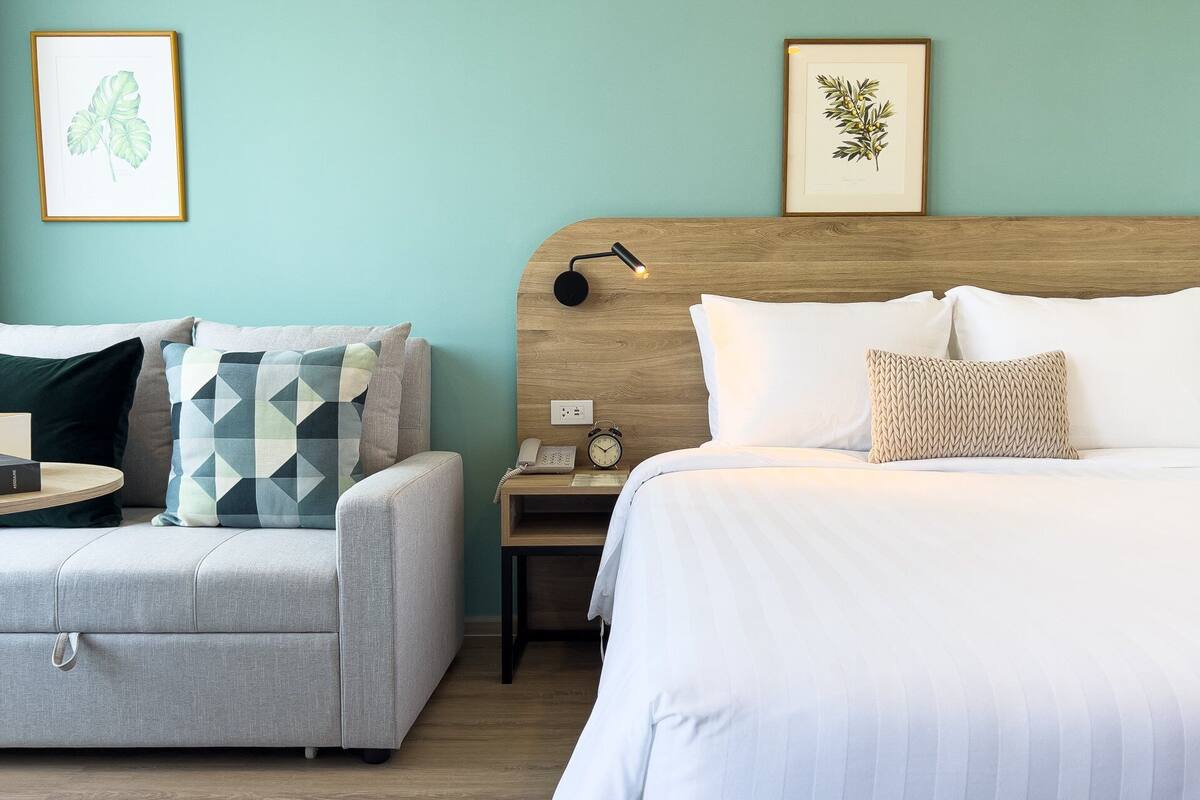 Habitación de hotel con cama, una silla, una mesita de noche con lámpara y dos grabados botánicos enmarcados en la pared.