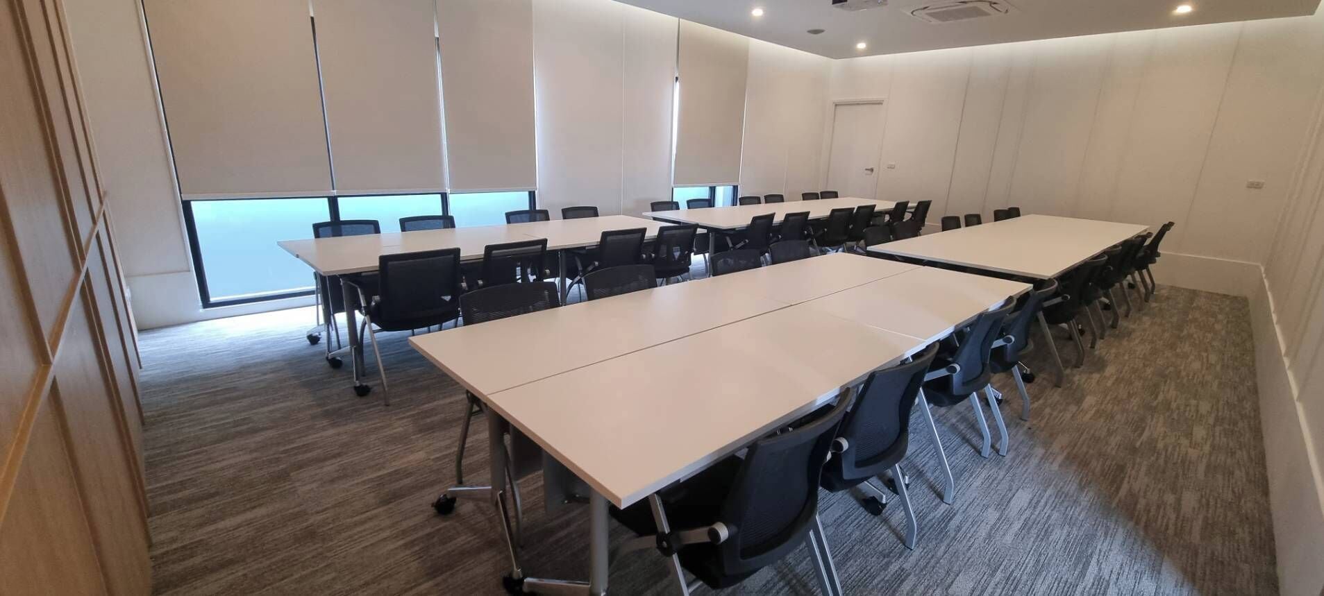 A conference room with multiple tables and chairs, carpeted flooring, and a large window providing natural light.