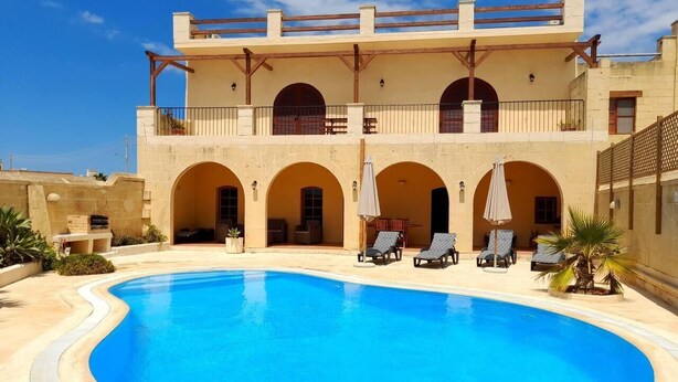 Zona de piscina con una pileta grande, sillas de descanso y un edificio con múltiples arcos y balcones.