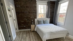 A bedroom with a brick wall, a large window, a bed with white bedding, and a wooden floor.