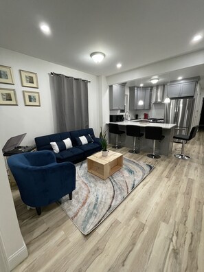 A modern living room with a blue sofa, a wooden coffee table, and a kitchen area with a stainless steel refrigerator and microwave. - Modern Luxury Apartment near NYC (Jersey City)
