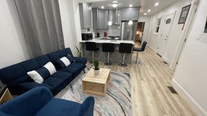 A modern living room with a blue sofa, a wooden coffee table, and a kitchen area with a stainless steel refrigerator and bar stools.