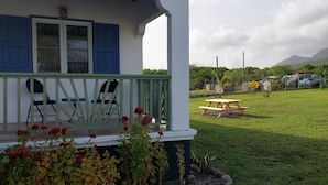 Una casa con un porche, una mesa y sillas. Hay un patio cubierto de césped con algunas plantas y una montaña al fondo.