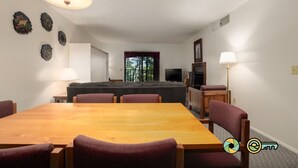 Een eetgedeelte met een houten tafel en zes stoelen, een bank, een televisie en een raam met uitzicht op bomen.