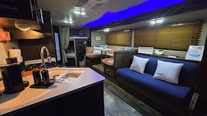 A compact kitchenette with a sink, stove, and storage cabinets. A blue sofa with white cushions is positioned near a window with blinds. - Enjoy a cozy space in a RV Camper w/hot tub, near beaches. (Lajas)