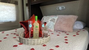 A bed with a white textured bedspread, a wicker basket containing a bottle of Gloria Margarita, two glasses, and red rose petals scattered on the bed. - Enjoy a cozy space in a RV Camper w/hot tub, near beaches. (Lajas)