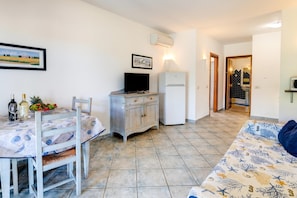 Une petite pièce bien éclairée, avec une table à manger, des chaises, un canapé, un téléviseur, un réfrigérateur et un coin cuisine visible à travers une porte ouverte.