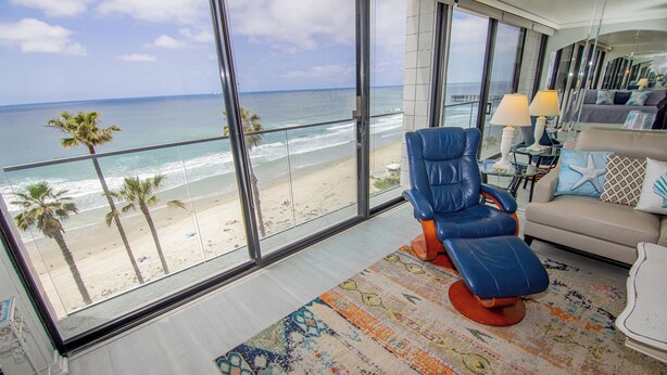 Una habitación moderna con un ventanal grande con vistas a la playa, un sillón de cuero azul, un sofá color beige y un tapete colorido.