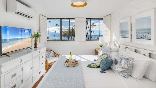 Un dormitorio moderno con una tele grande, una cama con vistas a la playa y un comedor con una bandeja de copas de vino.
