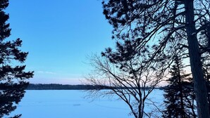 A snowy landscape with a clear view of a frozen lake, surrounded by trees and a building with a snow-covered roof. - Get Cozee Pequot Lakes, Lakefront on Mayo/Sibley (Pequot Lakes)