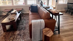 A living room with a brown leather couch, a wooden coffee table, and a patterned rug. There are large windows with a view of a deck and trees. - Get Cozee Pequot Lakes, Lakefront on Mayo/Sibley (Pequot Lakes)