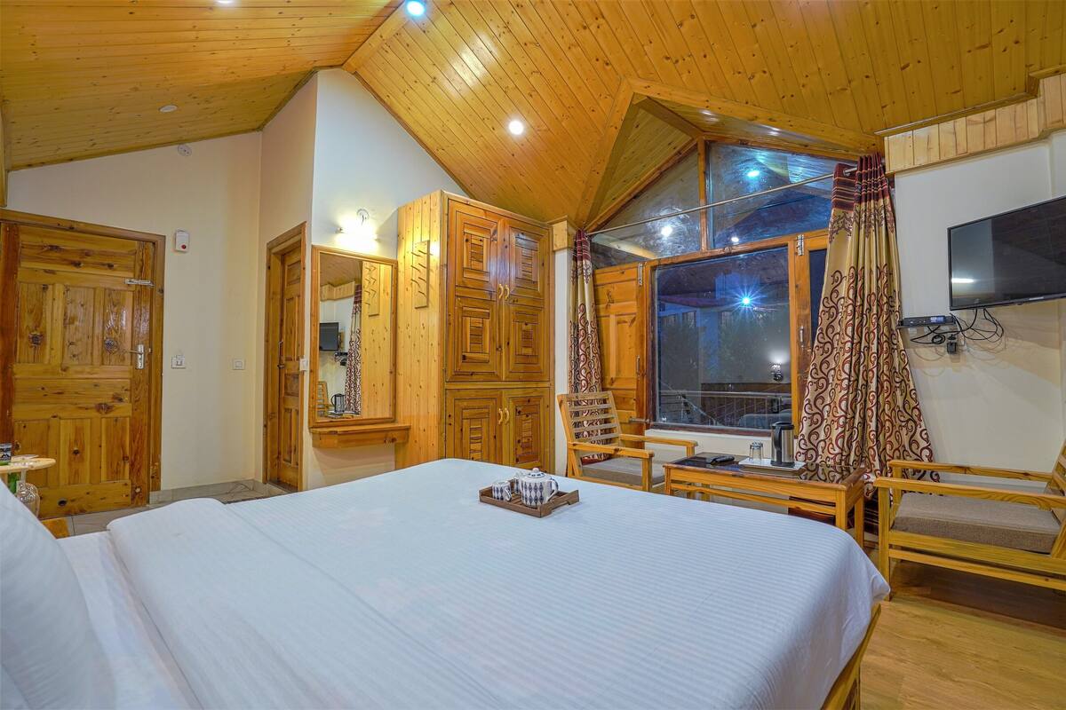 A wooden-paneled bedroom with a large bed, a chair, a TV, and a window with curtains.