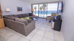 Ein Wohnzimmer mit einem großen grauen Ecksofa, einem kleinen runden Couchtisch, einem Beistelltisch mit Lampe, einem Bücherregal und einem an der Wand montierten Flachbildfernseher.