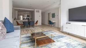 Ein modernes Wohnzimmer mit Sofa, Couchtisch und TV-Schrank. Im Hintergrund befindet sich ein Essbereich mit Stühlen und eine Küche.