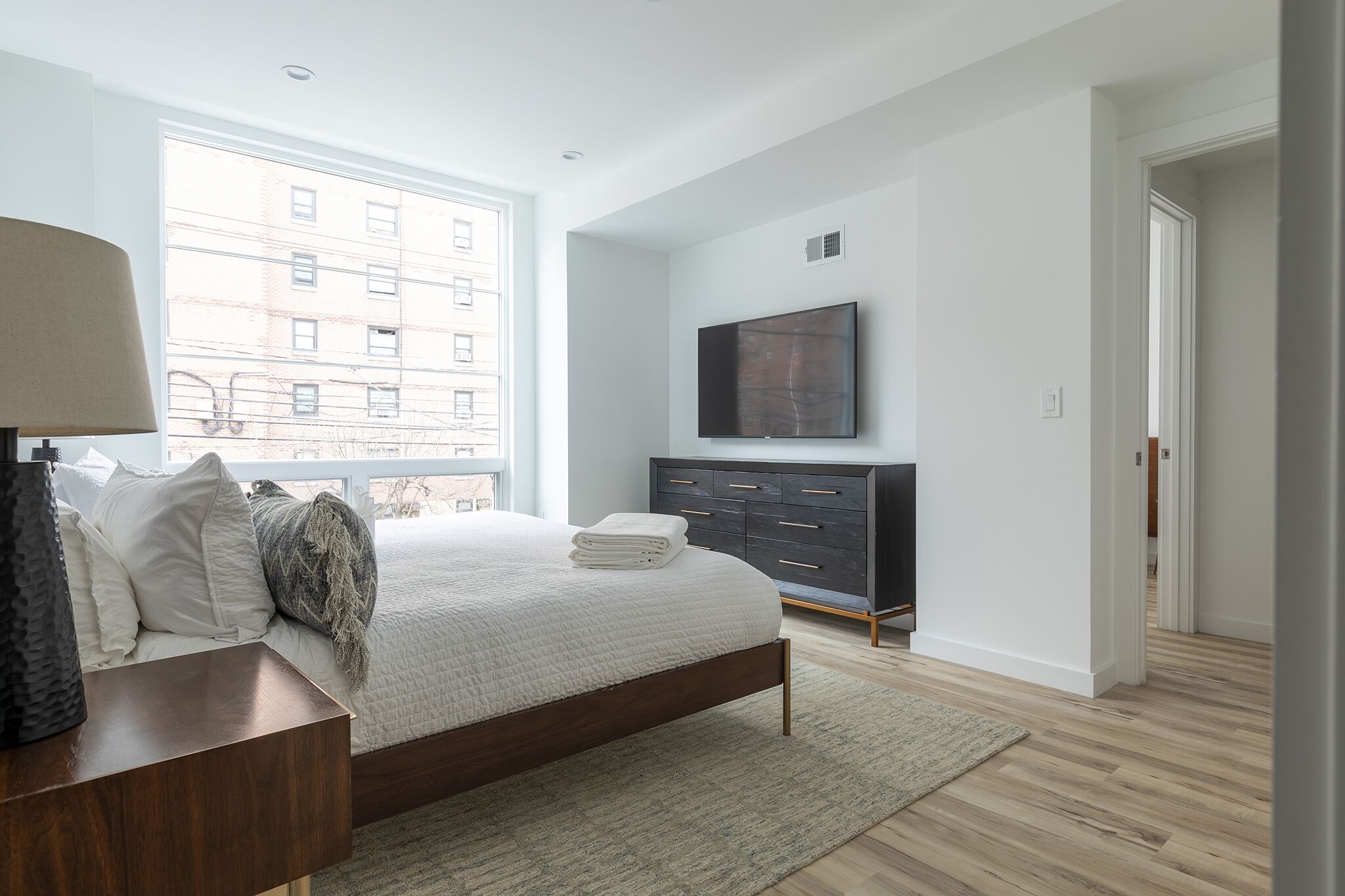 A modern bedroom with a large bed, a dark dresser, a flat-screen TV, and a view of a building through a large window.