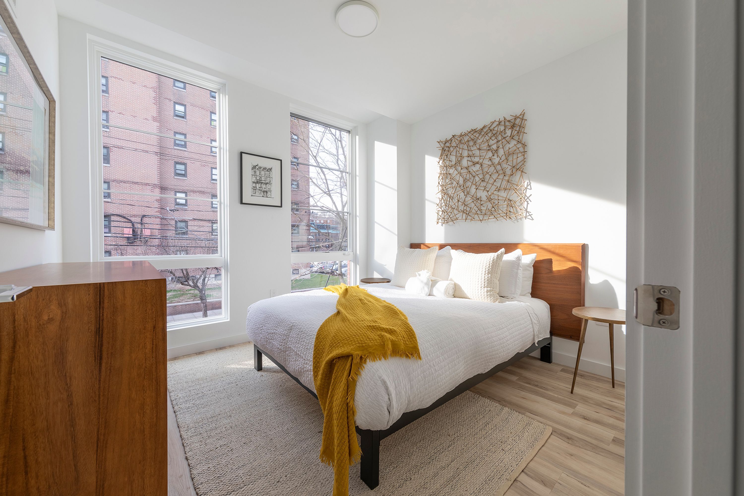 A modern bedroom with a large bed, a wooden dresser, and a view of a cityscape through large windows.
