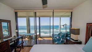 A hotel room with a large bed, a desk, a chair, a table, and a view of the ocean through large windows.