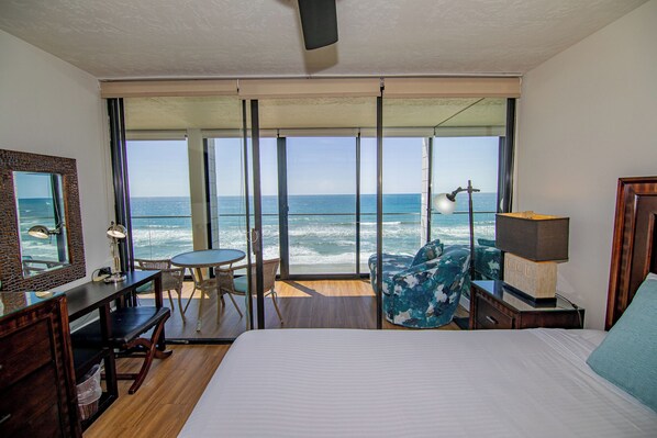 A hotel room with a large bed, a desk, a chair, a table, and a view of the ocean through large windows.