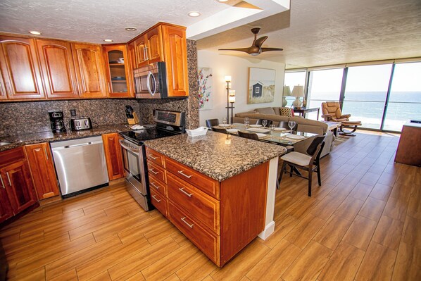 A modern kitchen with granite countertops, wooden cabinets, and stainless steel appliances. There is a dining area with a large window overlooking the ocean.