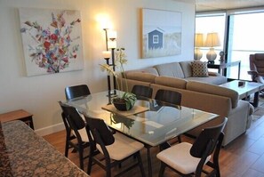 A modern living room with a glass dining table, beige sofa, and a large window offering a view.