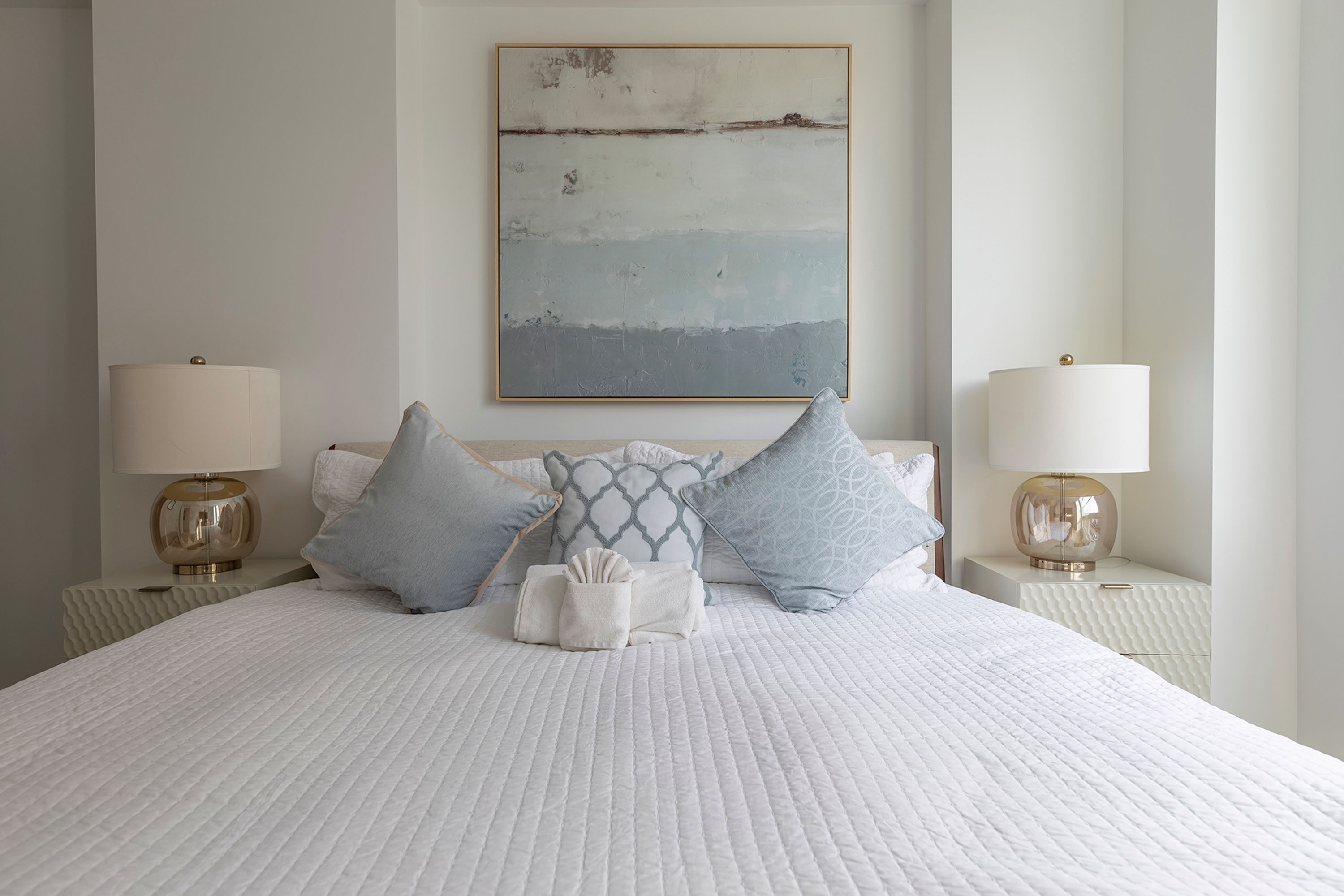 A neatly made bed with a quilted white cover, two bedside lamps, and a variety of pillows.