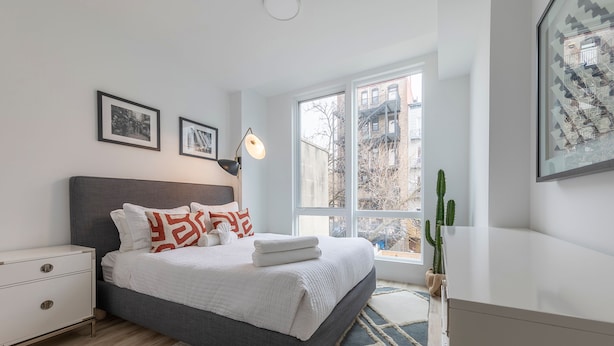 A modern bedroom with a large bed, bedside tables, a window with a view of buildings, and a cactus plant. - Sun-drenched 3BR Loft With Outdoor Space Near NYC (Hoboken)