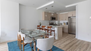 A modern kitchen with wooden cabinets, a stainless steel refrigerator, and a glass dining table with chairs. - Sun-drenched 3BR Loft With Outdoor Space Near NYC (Hoboken)