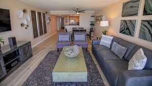 A spacious living room with a gray sofa, two armchairs, a coffee table, and a TV. A dining area is visible in the background with a kitchen. - ONE BEDROOM CONDO 609 (San Diego)
