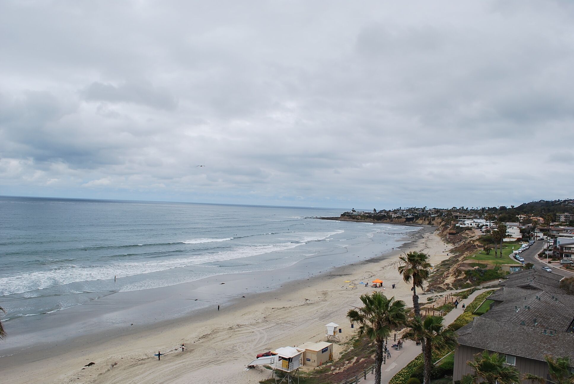 A coastal beach with a lifeguard tower, beach umbrellas, and a road with parked cars.