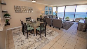 A spacious living area with a dining table, chairs, and a sofa. There is a ceiling fan, artwork on the walls, and a view of the ocean through large windows. - ONE BEDROOM CONDO 609 (San Diego)