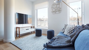 A modern living room with a large flat-screen TV, a sofa, ottomans, and a patterned rug. - Modern 3 BR Loft -shared Deck - gym - Mins to NYC (Hoboken)