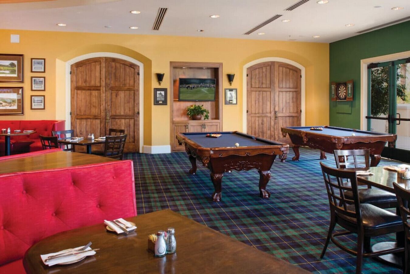 A billiards room with two pool tables, red seating, and framed pictures on the walls.