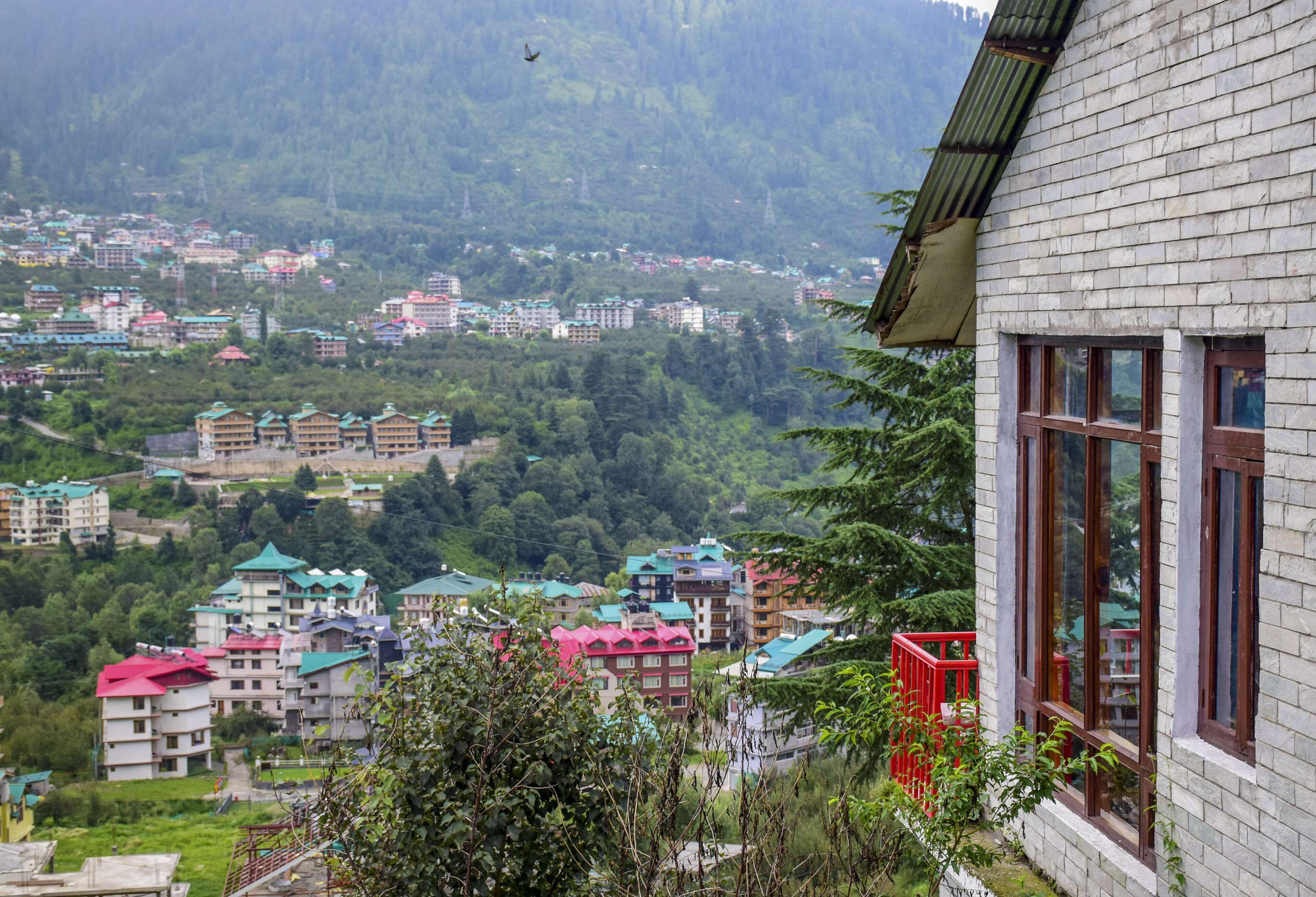 A view of a residential area with multiple buildings, some with colorful roofs, surrounded by lush greenery and hills in the background.