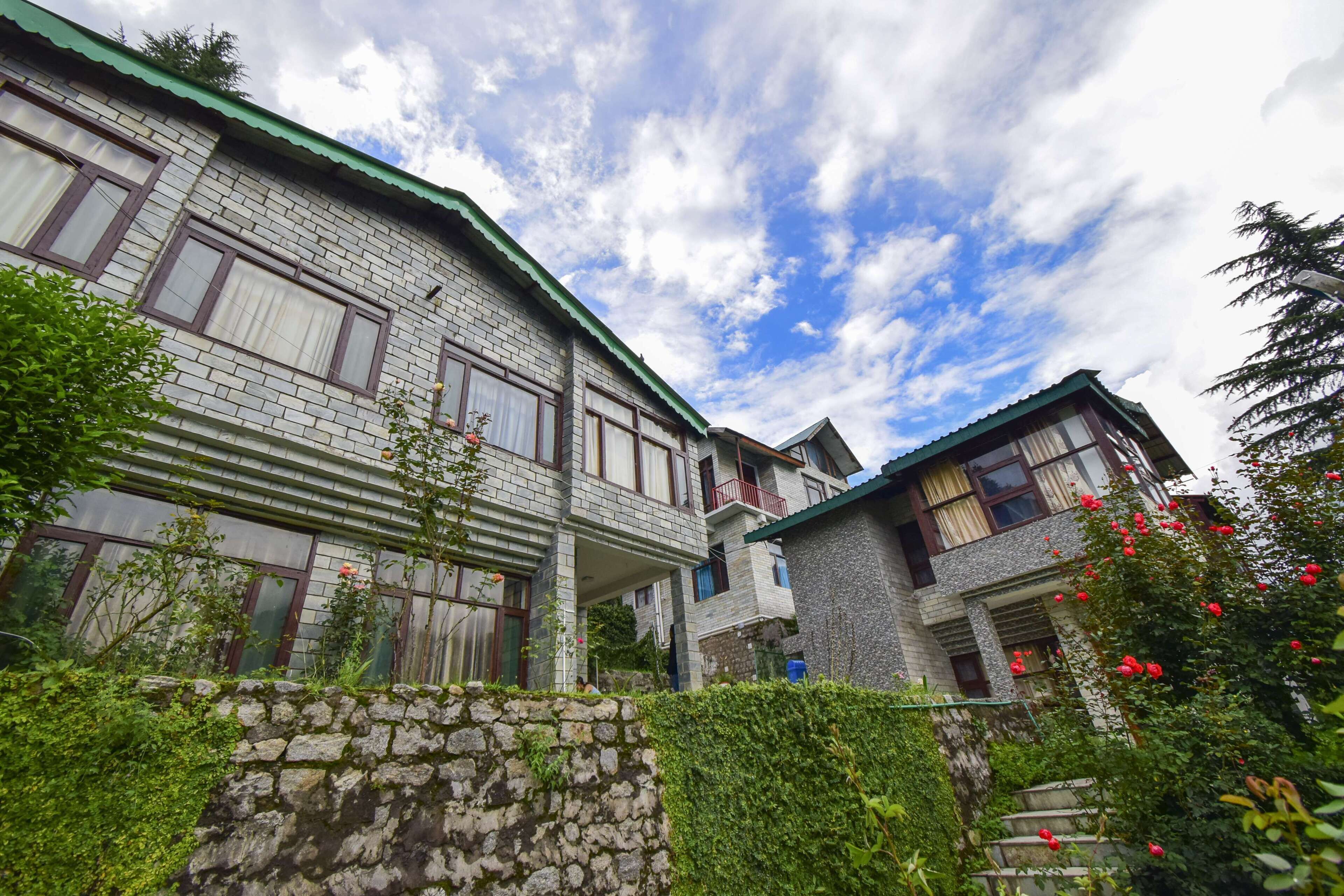 A traditional stone building with multiple windows and a green roof, surrounded by lush greenery and a stone wall.
