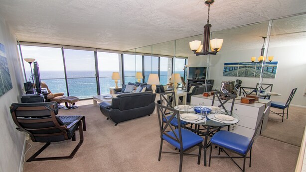 A modern living room with a view of the ocean, a dining area with blue chairs, and a kitchen area. - ONE BEDROOM CONDO 706 (San Diego)