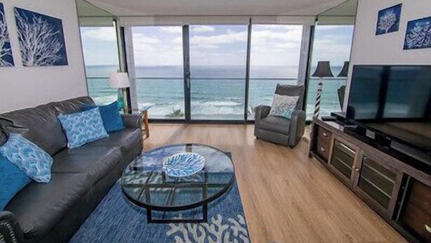 Uma sala de estar com uma grande janela com vista para o oceano, um sofá com almofadas azuis, uma mesa de centro de vidro e uma televisão sobre um móvel de madeira.