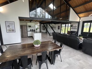A spacious kitchen with a central island, a dining area with a wooden table and black chairs, and a large window offering a view of the outdoors. - Large Home great for Families, Work, Groups, Teams (Rukuhia)