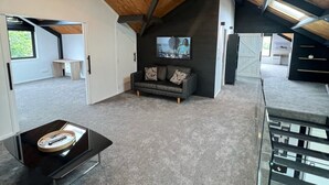 Un loft espacioso con techo alto, vigas a la vista, piso alfombrado en gris, una mesa de centro negra, un sofá gris con cojines estampados, un televisor de pantalla plana montado en la pared y una escalera con barandas de vidrio.