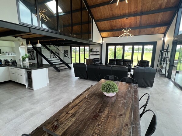 A spacious modern kitchen with a central island, a dining area with a wooden table, and a living area with black leather sofas. - Large Home great for Families, Work, Groups, Teams (Rukuhia)