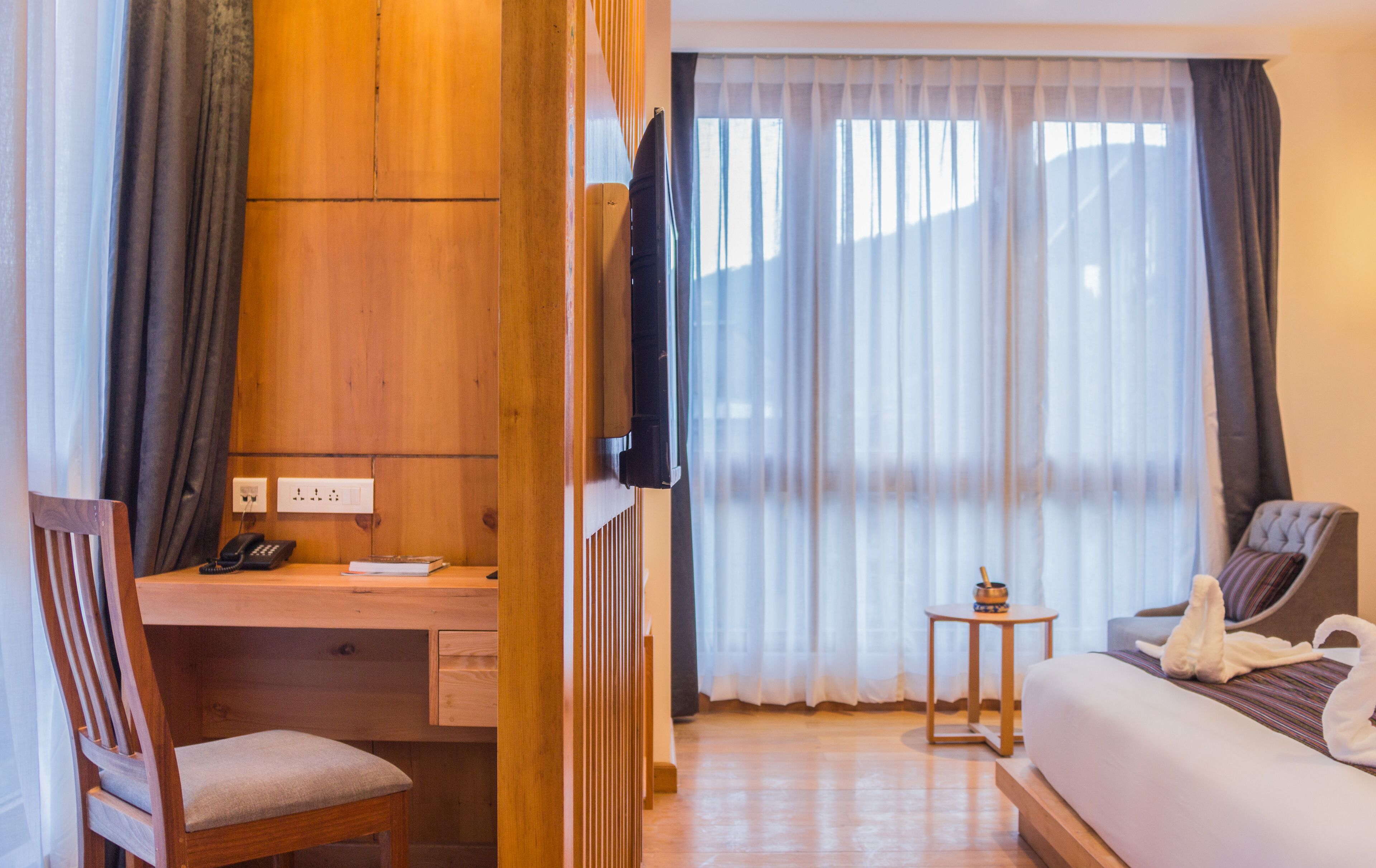 A hotel room with a wooden desk, a chair, a bed with a swan-shaped pillow, a TV, and a window with sheer curtains.