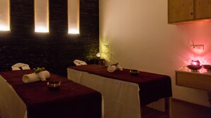 Un cuarto de spa con dos camillas de masaje, cada una cubierta con una sábana blanca y una manta roja. Hay toallas y un tazón sobre las camillas. La habitación tiene una pared de piedra oscura con tres ventanas iluminadas. Un mueble de madera y una pequeña mesa con una lámpara roja están a la derecha.