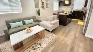 Ein modernes Wohnzimmer mit grauem Sofa, einem weißen Sessel und einem Couchtisch. Es gibt eine Küchenzeile mit dunklen Schränken und einem gelben Teppich.