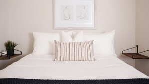 Un lit bien fait, avec du linge de lit blanc et des oreillers à rayures, une œuvre d’art encadrée au-dessus, et une petite plante posée sur une table de chevet.