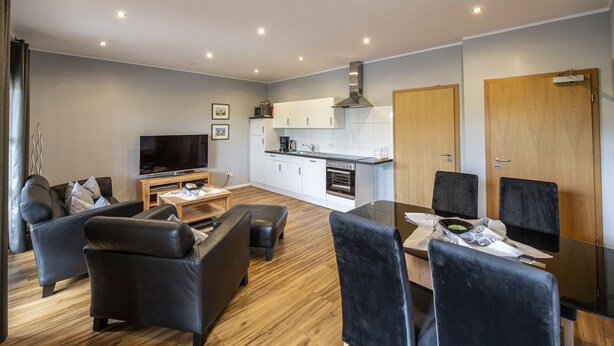 A modern living room with a sofa, ottoman, and a flat-screen TV. There is a dining area with a glass table and chairs. The kitchen is visible with white cabinets and a stove. - FerBeautiful apartment close to the Netherlands (Ahaus)