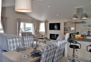 A modern living room with a sofa set, a dining table set for four, a kitchen area with a TV, and a view of the outdoors. - Sirona Lodge. Percy Woods (Percy Wood Country Park, Swarland)